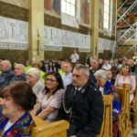 Na zdjęciu widzimy ludzi siedzących w kościelnych ławkach. Ściany ozdobione są freskami i tekstami w jasnych tonach. W tle widać rusztowanie, co sugeruje remont.