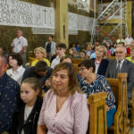 Grupa ludzi siedzi w ławkach w kościele. W tle widać dekoracje ścienne z napisami. Tło jest w kolorach brązu i beżu.