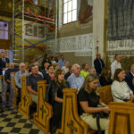 Grupa osób siedzi w kościelnych ławkach, wnętrze z malunkami na ścianach. Kolory są ciepłe, przewaga brązu i beżu. Z boku stoi kobieta z wózkiem.