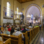 Wnętrze kościoła z wiernymi na ławkach, jasne witraże i freski na ścianach. Żyrandole oświetlają pomieszczenie. Msza święta trwa.