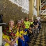 Grupa osób siedzi w budynku z zabytkowymi freskami. Trzy starsze kobiety noszą kolorowe, ludowe stroje. Posadzka jest w czarno-białą kratkę.