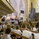 Grupa ludzi siedzi w kościele z jasnym wystrojem wnętrza. Na ołtarzu jest kapłan w zielonej szacie. Po lewej stronie widać orkiestrę z instrumentami dętymi.