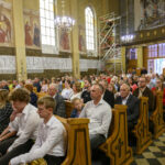 Grupa osób siedzi w kościele z drewnianymi ławkami, muralami i organami w tle. Dominują kolory brązowy i złoty. Na ścianach widoczne są freski i teksty.