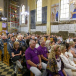 Wnętrze kościoła, ludzie siedzą w ławkach. Ściany zdobią freski, a światło pada przez duże okna. Tło jasne, sufit z żyrandolem.