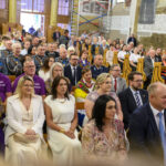 Grupa ludzi siedzących w kościele, większość w eleganckich ubraniach. Dominują kolory fioletowy, biały i szary. W tle widać dekoracje ścienne i muzyków.