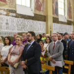 Grupa ludzi stoi w kościele z freskami na ścianach. Tło jest jasne z białymi i złotymi odcieniami. Wszyscy są odświętnie ubrani.