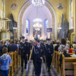 Grupa mężczyzn w mundurach maszeruje w kościele z witrażami. Podłoga w szachownicę, złote detale architektury. Eleganckie żyrandole świecą nad tłumem.