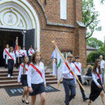 Grupa młodzieży w białych koszulach z czerwonymi szarfami wychodzi z ceglanego kościoła. Trzymają flagi i maszerują po bruku. Tło stanowi zielone drzewo i ceglana fasada.