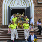 Na zdjęciu grupa ludzi wychodzi z ceglanego kościoła. Noszą kolorowe stroje, trzymają dekorację z kwiatów. W centrum chłopiec z dużym słonecznikiem.