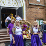 Grupa osób w barwnych, fioletowych strojach wychodzi z kościoła z ceglanymi ścianami. Tło stanowią otwarte drzwi i obraz Madonny nad wejściem. Przed nimi uformowana dekoracja z kwiatów i zbóż.