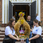 Ludzie trzymają wieńce z kłosów i słoneczników przed kościołem. W tle otwarte drzwi i ceglana ściana. Kolory naturalne.