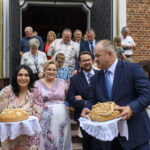 Grupa ludzi wychodzi z kościoła, trzymając tradycyjne chleby. Budynek z cegły w tle. Kobiety w kolorowych strojach.