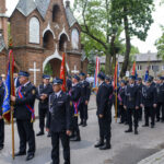 Parada strażaków w uniformach przed ceglanym kościołem. Trzymają flagi w barwach niebieskich i czerwonych. W tle drzewa i zgromadzony tłum.