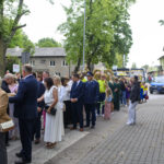 Grupa ludzi stoi w kolejce na ulicy, otoczona drzewami. W tle niebieski policyjny pojazd na tle budynków. Przeważają zielone i szare kolory.