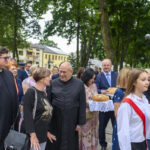 Na zdjęciu jest zbieg, grupa osób, m.in. duchowni i dzieci w strojach galowych, na tle zielonego parku. Dzieci niosą chleb na ozdobnych tacach. W tle widoczne są drzewa i budynki.