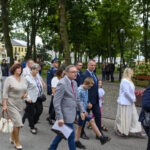 Grupa ludzi spaceruje alejką w parku, ubrani formalnie. W tle drzewa i budynki. Atmosfera jest spokojna i elegancka.