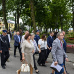 Grupa ludzi, w tym strażak, spaceruje po parku. W tle widać drzewa i budynki. Dominuje zieleń i stonowane kolory ubrań.