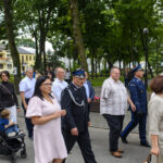 Grupa ludzi, w tym policjant, maszeruje alejką w parku. Otaczają ich wysokie drzewa, a w tle widoczne są budynki. Kolory są żywe, atmosfera spokojna.