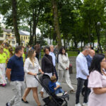 Grupa ludzi idzie w parku, w tle zielone drzewa i budynki. Kolorowe stroje uczestników. Przód obrazu zajmuje dziecko w wózku.