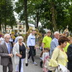 Grupa ludzi uczestniczy w uroczystym pochodzie na tle zielonych drzew i budynków. Stroje w jasnych kolorach, w tym odcienie zieleni i fioletu. W tle widoczna dekoracja z kwiatów i zbóż.
