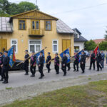 Grupa osób w mundurach niesie flagi podczas marszu ulicą. W tle żółty budynek i samochód. Dzień jest pochmurny.