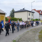 Parada na ulicy z uczestnikami w mundurach i sztandarami. Tło to budynki w stonowanych kolorach. Ludzie idą w zorganizowanej grupie.