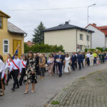 Grupa ludzi w eleganckich strojach maszeruje ulicą. Niektórzy niosą flagi, a w tle widoczne są domy. Pogoda jest pochmurna.