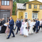 Grupa elegancko ubranych osób idzie ulicą. W tle ceglana kamienica i żółty dom. Atmosfera uroczystości formalnej.