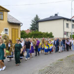 Grupa ludzi idzie ulicą podczas procesji dożynkowej. Ubrania w kolorach zieleni i fioletu, a tło to żółty budynek i krzewy. Widać ozdobne wieńce z kwiatów i zbóż.