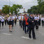 Orkiestra maszeruje ulicą, grając na instrumentach dętych. Uczestnicy ubrani są w białe koszule i ciemne spodnie. Tło stanowią drzewa i budynki.