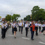 Orkiestra maszeruje ulicą w białych i czarnych mundurach. W tle zielone drzewa i billboardy. Dzień pochmurny.
