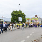 Grupa ludzi maszeruje ulicą, ubrani w różne kolory. W tle zielone drzewo i budynek z reklamami. Niebo jest zachmurzone.