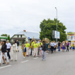 Grupa ludzi maszeruje ulicą podczas procesji, wielu ubranych kolorowo. W tle zielone drzewa i reklamy. Atmosfera jest świąteczna.