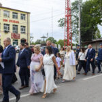 Grupa elegancko ubranych ludzi idzie ulicą. W tle żółty budynek z napisem Dom Strażaka. Po prawej wysokie drzewa i wieża.