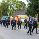 Parada strażaków w mundurach na ulicy, niosących flagi. Tło wypełnia zielona roślinność i budynki. Niebo zachmurzone.