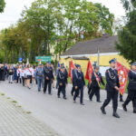 Parada strażaków i mieszkańców w mundurach na ulicy. Zielone drzewa, żółty budynek w tle. Flagi czerwone i niebieskie.