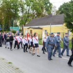Grupa ludzi w paradzie na ulicy, niektórzy w mundurach z flagami. W tle żółty budynek i zielona roślinność. Długa procesja z tablicą informacyjną.