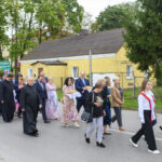 Grupa ludzi maszeruje ulicą obok żółtego budynku, niesie flagę Polski. W tle drzewa i zielony znak drogowy. Atmosfera uroczysta.