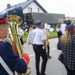 Grupa muzyków maszeruje ulicą w strojach paradnych. W tle widać budynek i ciężarówkę. Dominują kolory niebieski, biel i czerwień.
