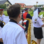 Orkiestra dęta na ulicy. Członkowie w białych koszulach grają na instrumentach. W tle domy i zielone tło.