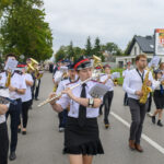 Grupa muzyczna w mundurach maszeruje ulicą. Dominuje biel i czerń, zielone drzewa w tle. Pogoda jest pochmurna.