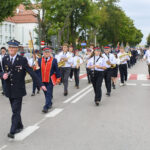 Orkiestra maszeruje ulicą w strojach galowych, grając instrumenty dęte. Tło to drzewa i budynek z czerwoną dachówką. Pogoda jest pochmurna.