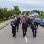 Strażacy w czarnych mundurach maszerują po ulicy. Niosą sztandary, a tło to zieleń i budynki. Obok parkują auta.