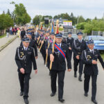 Grupa strażaków maszeruje w paradzie, ubrani na czarno z ozdobnymi naszywkami. W tle zielone drzewa i niebieski samochód. Pogoda jest pochmurna.