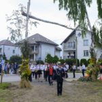 Parada orkiestry dętej w mundurach maszeruje drogą. Otoczenie to domy i drzewa, na trawie leżą dynie. Pogoda pochmurna.