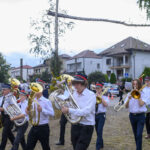 Orkiestra dęta w białych koszulach idzie po trawie, trzymając instrumenty. W tle domy i błękitne niebo.