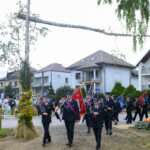 Grupa strażaków maszeruje z czerwonymi flagami, ubrani w ciemne mundury. Tło stanowią domy i zielone drzewa. Nad nimi drewniana konstrukcja z dekoracjami z kwiatów i traw.