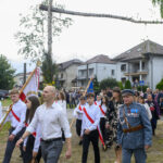Grupa osób w białych koszulach maszeruje z flagami. W tle widać budynki mieszkalne i drzewo. Starszy mężczyzna w mundurze prowadzi pochód.