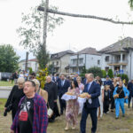 Grupa ludzi w eleganckich ubraniach maszeruje, niosąc chleb. W tle biały dom i drzewo ozdobione kwiatami. Dzień jest pochmurny.