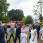 Grupa osób elegancko ubranych przechodzi obok czerwonego wozu strażackiego na tle drzew. W tle widoczny jasny niebieski niebo i zielone drzewa. Po prawej stronie znajduje się drewniany krzyż udekorowany kwiatami.